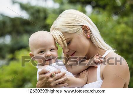 A beautiful young mom cuddling her baby daughter who is posing for the camera while enjoying quality time outdoors in a park on a warm summer day; Edmonton, Alberta, Canada View Large Photo Image Stock Photograph - A beautiful young mom cuddling her baby daughter who is posing for the camera while enjoying quality time outdoors in a park on a warm summer day; Edmonton, Alberta, Canada. Fotosearch