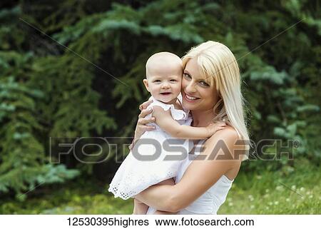 Stock Photograph - A beautiful young mom cuddling her baby daughter who is posing for the camera while enjoying quality time outdoors in a park on a warm summer day; Edmonton, Alberta, Canada. Fotosearch