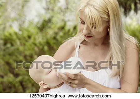 Stock Photograph - A beautiful young mom feeding her sleepy baby daughter while enjoying quality time outdoors in a park on a warm summer day; Edmonton, Alberta, Canada. Fotosearch