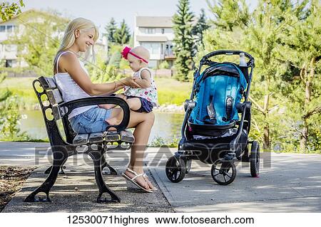 Stock Photograph - A beautiful young mother holding her baby daughter on her knees while resting on a park bench beside the lake after walking with a stroller on a warm sunny day; Edmonton, Alberta, Canada. Fotosearch