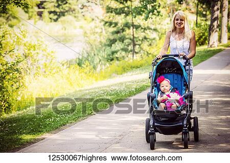 Stock Photograph - A beautiful young mother taking her baby daughter out for a walk using a stroller in a park with a lake on a warm sunny day; Edmonton, Alberta, Canada. Fotosearch