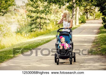 Stock Photograph - A beautiful young mother taking her baby daughter out for a walk using a stroller in a park with a lake on a warm sunny day and talking on her cell phone; Edmonton, Alberta, Canada. Fotosearch