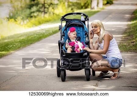 Stock Photograph - A beautiful young mother taking her baby daughter out for a walk using a stroller in a park with a lake on a warm sunny day and stopping to take a call on her cell phone; Edmonton, Alberta, Canada. Fotosearch