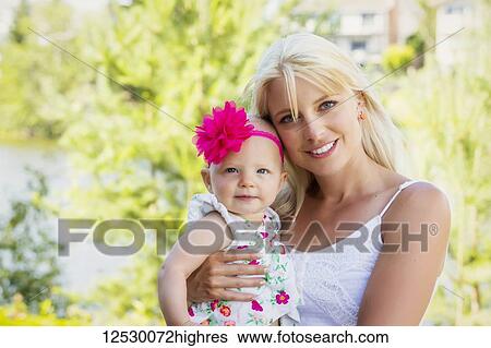 Stock Photograph - A beautiful young mother with long blonde hair enjoying quality time with her cute baby daughter in a city park on a summer day and posing for the camera; Edmonton, Alberta, Canada. Fotosearch