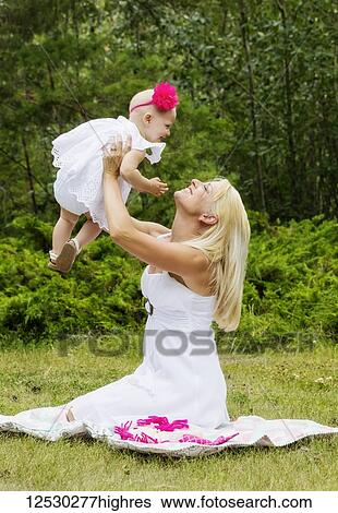 A beautiful young mother with long blonde hair enjoying quality time with her cute baby daughter and tossing her in the air while sitting on the grass in a city park during a summer day; Edmonton, Alberta, Canada View Large Photo Image Stock Photograph - A beautiful young mother with long blonde hair enjoying quality time with her cute baby daughter and tossing her in the air while sitting on the grass in a city park during a summer day; Edmonton, Alberta, Canada. Fotosearch