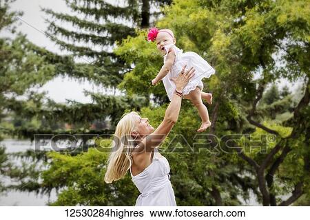 A beautiful young mother with long blonde hair enjoying quality time with her cute baby daughter and tossing her in the air in a city park on a summer day; Edmonton, Alberta, Canada View Large Photo Image Stock Photograph - A beautiful young mother with long blonde hair enjoying quality time with her cute baby daughter and tossing her in the air in a city park on a summer day; Edmonton, Alberta, Canada. Fotosearch