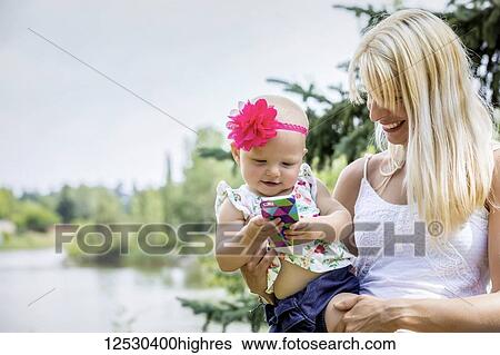 Stock Photograph - A beautiful young mother with long blonde hair enjoying quality time with her cute baby daughter who is amused with a smart phone in a city park on a summer day; Edmonton, Alberta, Canada. Fotosearch