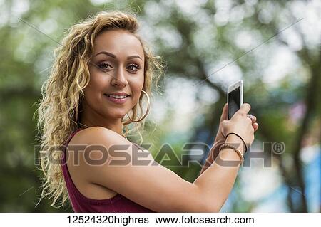 Stock Photograph - A beautiful young woman stops and poses for the camera while texting on her smart phone; Edmonton, Alberta, Canada. Fotosearch
