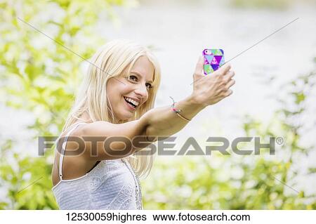 Stock Photograph - A beautiful young woman taking a self-portrait while enjoying her alone time in a park with a manmade lake; Edmonton, Alberta, Canada. Fotosearch