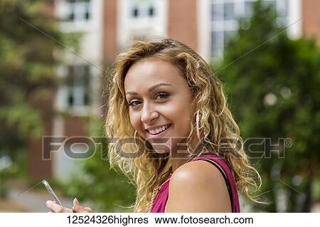 Stock Photograph - A beautiful young woman texting on her smart phone on a university campus and pausing to look at the camera; Edmonton, Alberta, Canada. Fotosearch