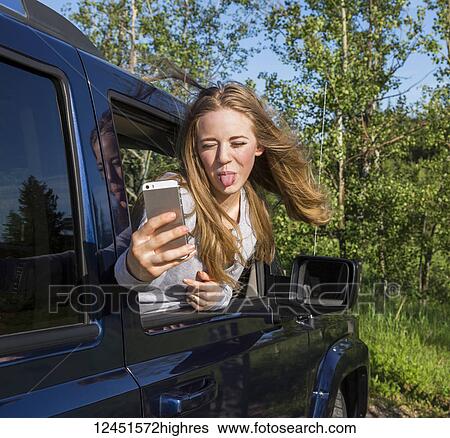A beautiful young woman with long blond hair looking out a vehicle window and taking a silly self-portrait with her smart phone while sticking out her tongue to the camera; Edmonton, Alberta, Canada View Large Photo Image Stock Photograph - A beautiful young woman with long blond hair looking out a vehicle window and taking a silly self-portrait with her smart phone while sticking out her tongue to the camera; Edmonton, Alberta, Canada. Fotosearch