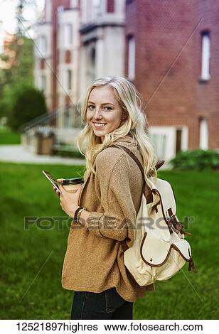 Stock Photograph - A beautiful young woman with long blond hair holding a coffee cup and texting on her smart phone while walking in a university campus stops to pose for the camera; Edmonton, Alberta, Canada. Fotosearch