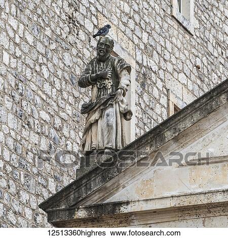 Stock Photograph - A bird perched on the head of a statue beside a stone wall of a building; Perast, Kotor Municipality, Monte. Fotosearch