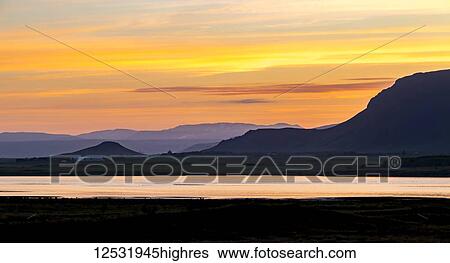 Stock Photograph - A colourful view at sunset over the water as dusk creates a silhouette of the hills of Selfoss in Western Iceland close to midnight, where the sun doesn't fully set around summer solstice; Selfoss, Iceland. Fotosearch