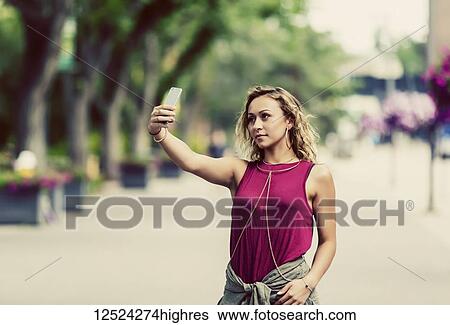 Stock Photograph - A conceptual shot of a young woman taking a self-portrait on a smart phone while down the street by herself near a university campus; Edmonton, Alberta, Canada. Fotosearch