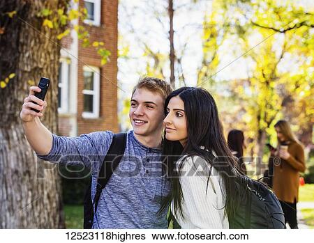 A couple who are university students posing for a self-portrait with a smart phone with a small group of friends in the background; Edmonton, Alberta, Canada View Large Photo Image Stock Photograph - A couple who are university students posing for a self-portrait with a smart phone with a small group of friends in the background; Edmonton, Alberta, Canada. Fotosearch