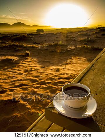 Stock Photograph - A cup of coffee sits on a wooden table with a view of the sunrise over the desert; Sossusvlei, Hardap Region, Namibia. Fotosearch