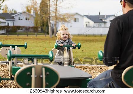 Stock Photograph - A cute young girl playing on a seesaw with her dad in a playground during the fall season; Spruce Grove, Alberta, Canada. Fotosearch
