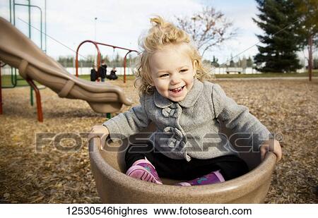 Stock Photograph - A cute young girl spinning in a saucer on a playground during the fall season; Spruce Grove, Alberta, Canada. Fotosearch