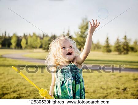 Stock Photograph - A cute young girl with blond hair blowing a bubble and trying to catch it in a park on a warm fall day; Spruce. Fotosearch