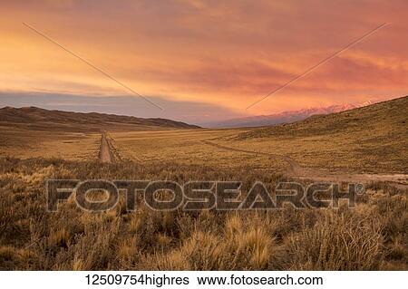 Stock Photograph - A dirt road leads the eye towards the horizon through bare desert hills. A snow-capped mountain range is visible in the distance. The scene and the clouds are lit by a red early sunrise; Potrerillos, Mendoza, Argentina. Fotosearch