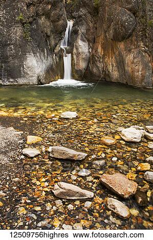 Stock Photograph - A double waterfall between dark rock walls with the foreground of colourful small rocks and pebbles; Calgary, Alberta, Canada. Fotosearch