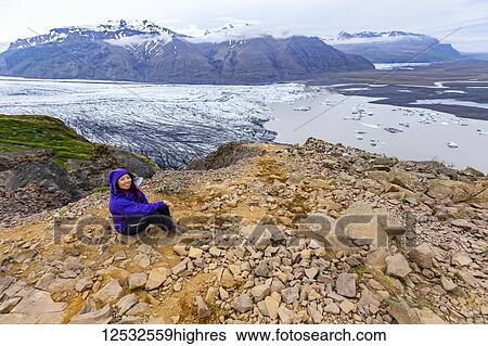 Stock Photograph - A female hiker in warm clothing poses on a mountain top overlooking the glacier lake and valley below at Vatnajokull National Park; Iceland. Fotosearch