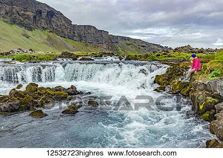 Stock Photograph - A female hiker poses for a photo on the edge of a waterfall; Iceland. Fotosearch