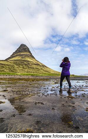 Stock Photograph - A female hiker stops on the beach at low tide to take a photo with an SLR camera in front of Kirkjufell Mountain in Snaefellsnes peninsula, Western Iceland; Iceland. Fotosearch