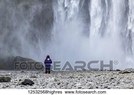 Stock Photograph - A female tourist looks towards the wall of water from Iceland's famous Skogafoss waterfall; Iceland. Fotosearch