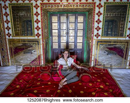 Stock Photograph - A female tourist sits on the luxurious red rug on the floor of the City Palace, Maharaja Sawai Mansingh II Museum, Mubarak Mahal; Jaipur, Rajasthan, India. Fotosearch