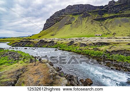 Stock Photograph - A fresh looking blue river runs along the edge of a farm property with volcanic mountains in the background; Iceland. Fotosearch