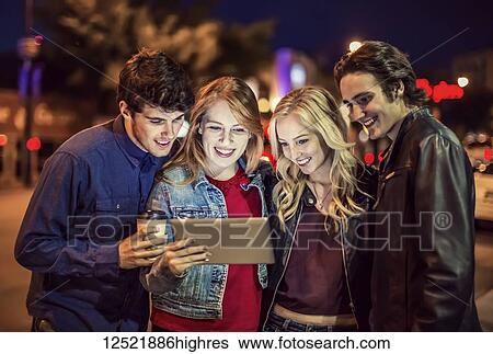 Stock Photograph - A group of four friends huddle together on a sidewalk looking at a tablet as the glow from the screen lights up their faces; Edmonton, Alberta, Canada. Fotosearch