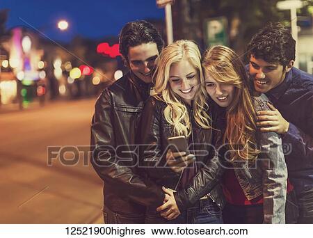 A group of four friends huddle together on a sidewalk looking at a smart phone as the glow from the screen lights up their faces; Edmonton, Alberta, Canada View Large Photo Image Stock Photograph - A group of four friends huddle together on a sidewalk looking at a smart phone as the glow from the screen lights up their faces; Edmonton, Alberta, Canada. Fotosearch