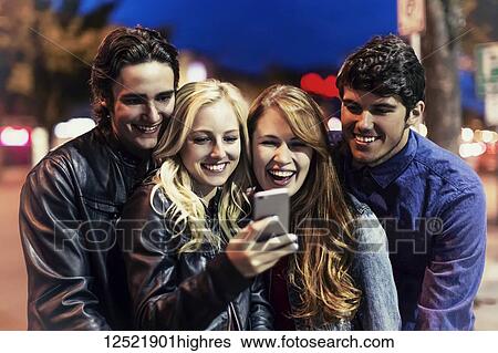 Stock Photograph - A group of four friends huddle together on a sidewalk looking at a smart phone as the glow from the screen lights up their faces; Edmonton, Alberta, Canada. Fotosearch