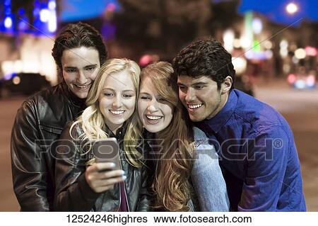 A group of four friends huddle together on a sidewalk looking at a smart phone as the glow from the screen lights up their faces; Edmonton, Alberta, Canada View Large Photo Image Stock Photograph - A group of four friends huddle together on a sidewalk looking at a smart phone as the glow from the screen lights up their faces; Edmonton, Alberta, Canada. Fotosearch