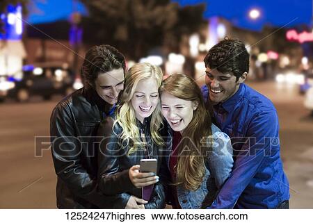 A group of four friends huddle together on a sidewalk looking at a smart phone and laughing as the glow from the screen lights up their faces; Edmonton, Alberta, Canada View Large Photo Image Stock Photograph - A group of four friends huddle together on a sidewalk looking at a smart phone and laughing as the glow from the screen lights up their faces; Edmonton, Alberta, Canada. Fotosearch