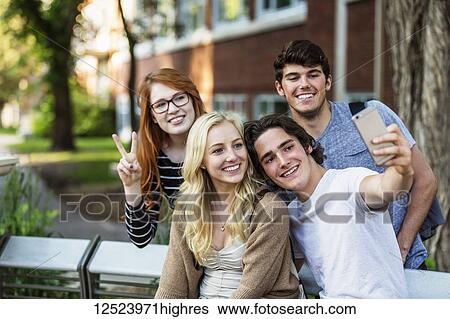 Stock Photograph - A group of four friends sitting on a bench taking a self-portrait with a smart phone on a university campus; Edmonton, Alberta, Canada. Fotosearch