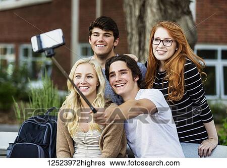 Stock Photograph - A group of four friends sitting on a bench taking a self-portrait with a smart phone using a selfie stick on a university campus; Edmonton, Alberta, Canada. Fotosearch