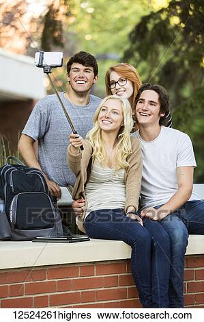 Stock Photograph - A group of four friends sitting on a bench taking a self-portrait with a smart phone using a selfie stick on a university campus; Edmonton, Alberta, Canada. Fotosearch