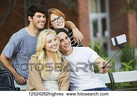 Stock Photograph - A group of four friends sitting on a bench taking a self-portrait with a smart phone using a selfie stick on a university campus; Edmonton, Alberta, Canada. Fotosearch
