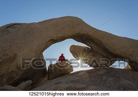 Stock Photograph - A hiker watching a sunrise under sandstone rock arch with sun star, Joshua Tree National Park; California, United States of America. Fotosearch