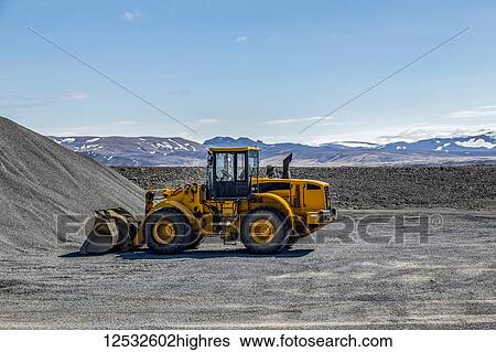 Stock Photograph - A large wheel loader heavy equipment machine is parked in front of a gravel pile in the mountain landscape; Iceland. Fotosearch