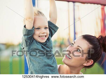 Stock Photograph - A little girl with blonde hair playing in in a playground on a warm fall day while her mom watches to keep her safe; Spruce Grove, Alberta, Canada. Fotosearch