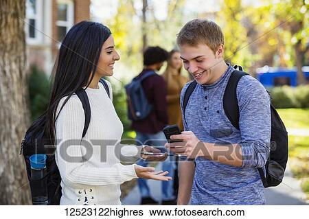 Stock Photograph - A male and female university student stand on a path looking at a smart phone together, laughing and talking together with a small group of fellow students in the background; Edmonton, Alberta, Canada. Fotosearch