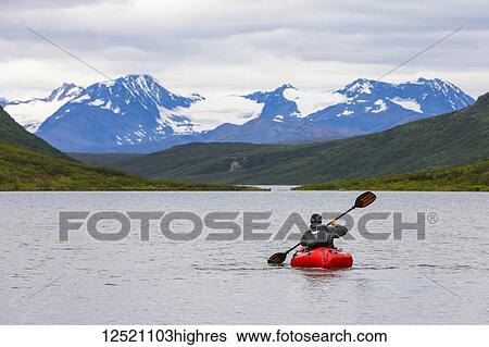 Stock Photograph - A man paddles a packraft across Landmark Gap Lake with the Alaska Range in the distance; Alaska, United States of America. Fotosearch