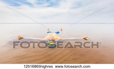 Stock Photograph - A man wearing a bathing suit floats in the shallow ocean water with his body outstretched and looking up to the sky; Prince Edward Island, Canada. Fotosearch