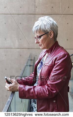 Stock Photograph - A mature woman in a red leather jacket stands at a railing using a smart phone; Vancouver, British Columbia, Canada. Fotosearch