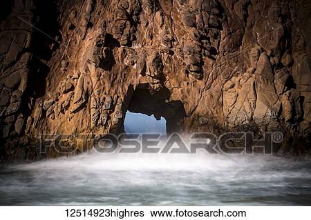 Stock Photograph - A natural arch in the rock formation along the coast with mist rising off the water; Big Sur, California, United States of America. Fotosearch