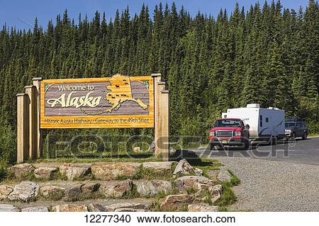 Stock Image - A pick up truck and RV camper parked at the Welcome to Alaska Highway Sign, Northern Yukon Territory, United States and Canadian Boarder, Summer. Fotosearch